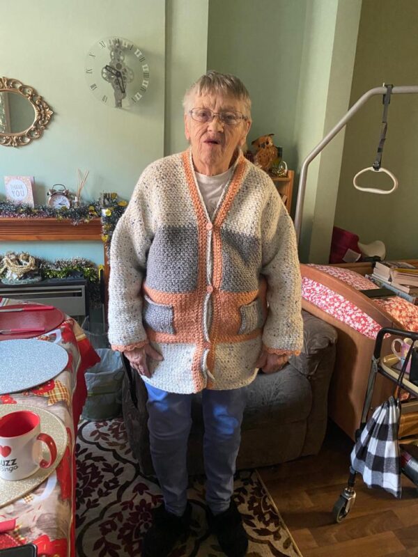 Old lade wearing a crochet cardigan in white, pink and grey horizontal panels. Sleeves are white with pink edging on the long sleeves. The neck and front edging are in pink and white. There are 3 multicoloured buttons in the front.